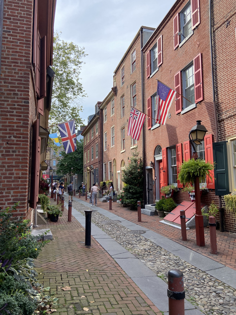 Elfreth's Alley. All of the homes are made of brick, with the closest home having red shutters. A home on the left has a British flag. A home on the right has a British-American flag and an American flag with thirteen stars.