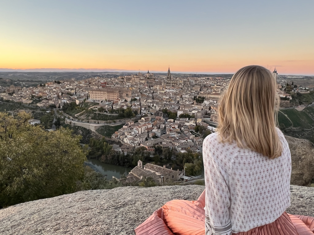 Stephanie (the author of this blog) sitting on the top of a cliff staring at the view of Toledo, Spain. A sunset is in the background of the photo. Stephanie is wearing a white long-sleeve shirt and a long, pink skirt.