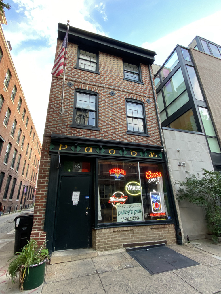 Paddy's Irish Pub - three-story brick building with American flag pole attached to it. Bottom floor windows have numerous neon signs on display. Above the windows is a sign in green and yellow that says "Paddy's".