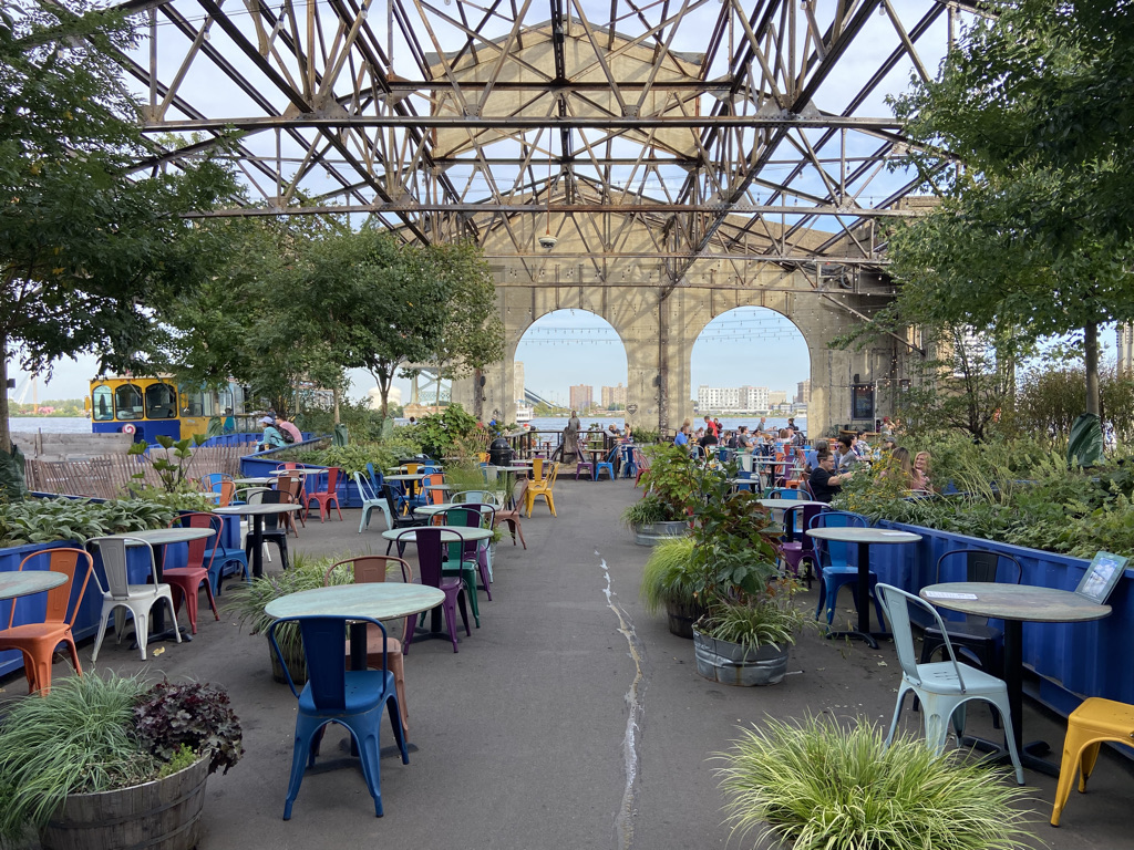 The beer garden in Cherry Street Pier. Outdoor space with greenery and colorful table and chair sets. Windows at end of pier overlook the river.