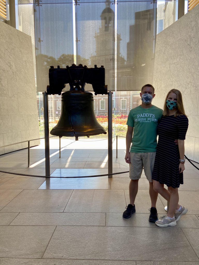 Stephanie (author of this blog post) wearing a mask, navy blue dress with yellow stripes, and off-white sneakers and her boyfriend Michael wearing a mask, a green Paddy's Pub t-shirt, khaki shorts, and black sneakers standing besides the Liberty Bell. 