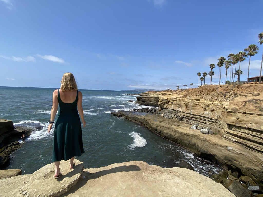 Stephanie (the author of this blog) in a mid-length forest green dress and sandals facing the ocean. She is standing on the edge of a cliff. On the right is another cliff and a row of palm trees.