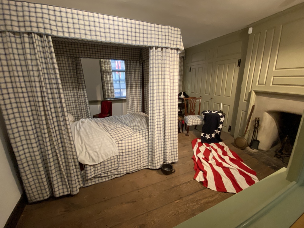 One of the bedrooms in Betsy Ross's house. The walls of the bedroom are an olive green with hardwood floors. The bedroom contains a bed with curtains around it. Both the curtains and comforter have a black and white checker pattern. Near the bed is a chair covered in the American flag with thirteen stripes.