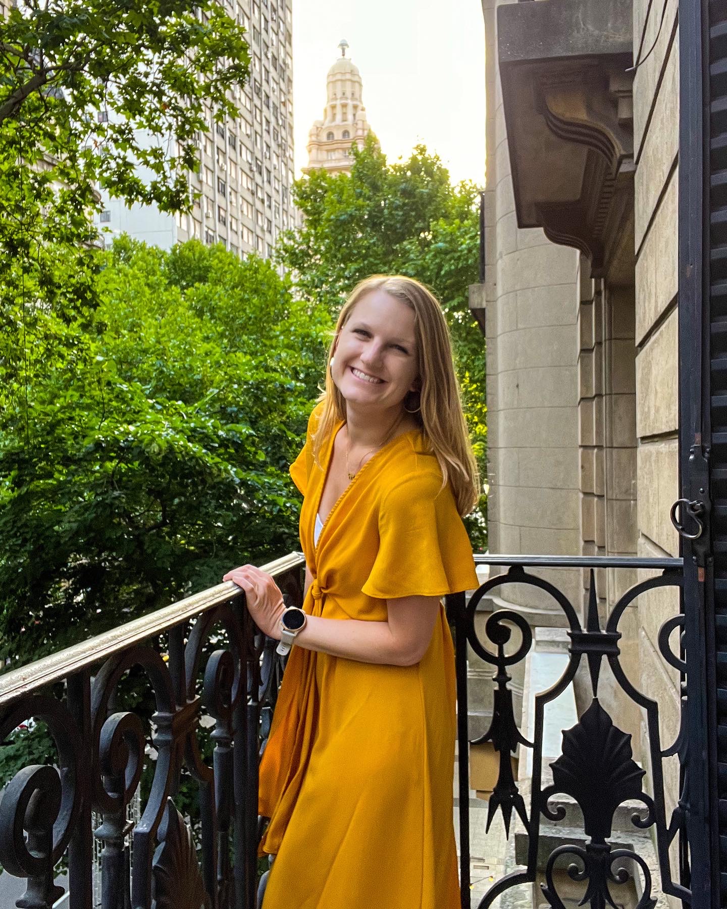 Stephanie (the author of this blog) is wearing a mid-length yellow dress and standing on a balcony. She is smiling at the camera. In the background are trees.