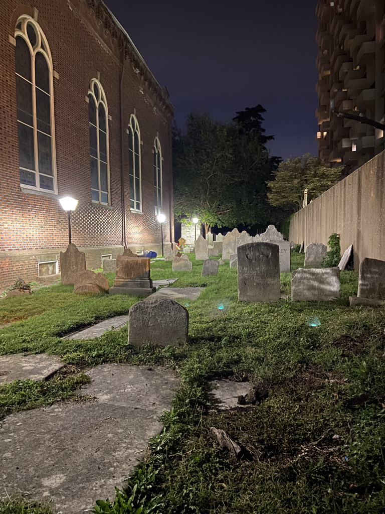 Narrow graveyard of 10-15 historic graves. The graveyard is between a church made of brick with tall windows and a fence.