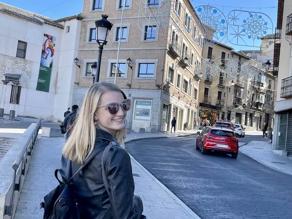 Stephanie (author of this post) is wearing sunglasses, a black leather coat, and a black backpack. She is standing on a sidewalk in Toledo, Spain with a red car, a road, and tan buildings in the background.