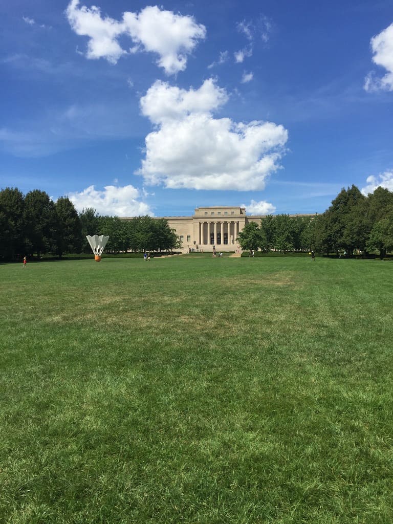 A large green lawn and the front of the Nelson-Atkins Museum of Art. A shuttlecock sculpture is standing to the left of the museum. The museum is also surrounded by trees with green leaves.