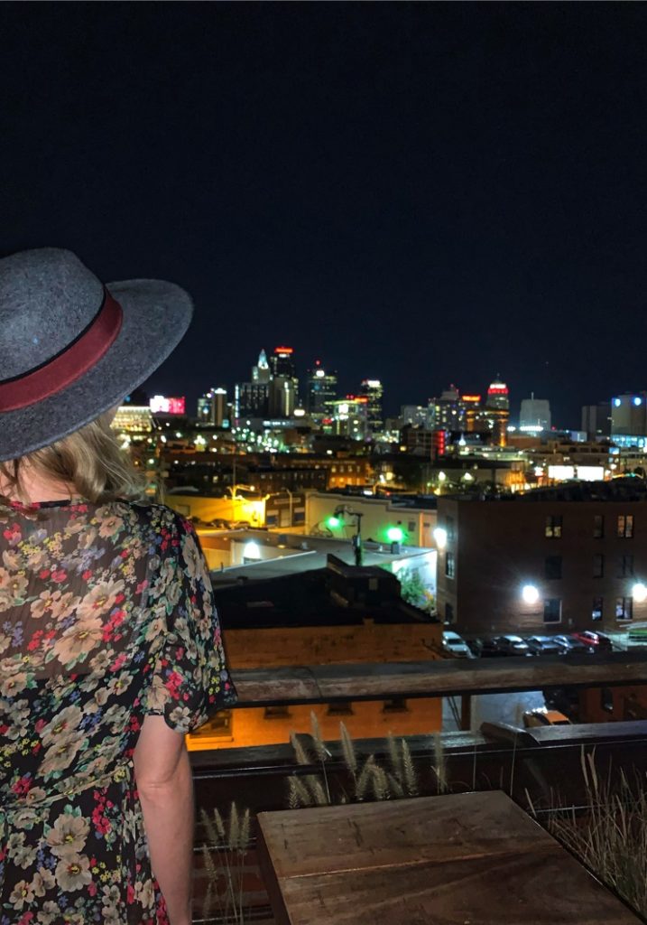 Stephanie (the author of this post) wearing a floral dress and gray hat and enjoying the view of downtown Kansas City at Percheron. The downtown Kansas City skyline is visible in the photo.