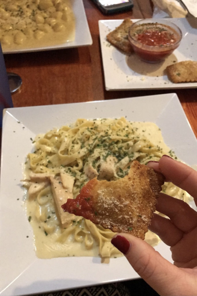 A few plates of food at Cupini's. The photographer is holding a toasted ravioli over a plate of chicken Alfredo. The other plates have more toasted raviolis and gnocci.