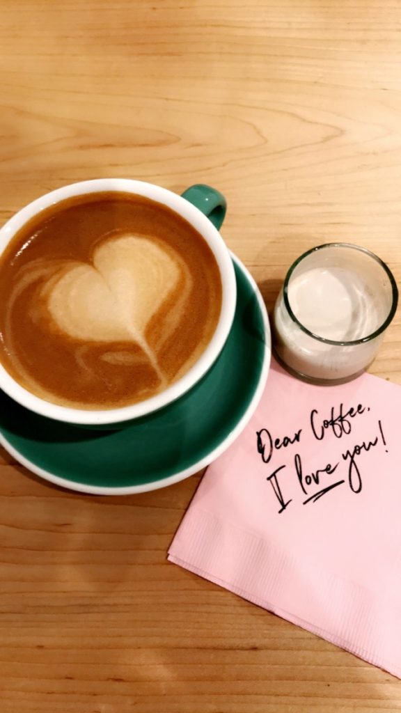 A cup of latte at Made in KC Cafe. The latte has a foam heart on top. There is a napkin next to the coffee cup that says "Dear Coffee, I love you!". These items are place on a bright wooden table.