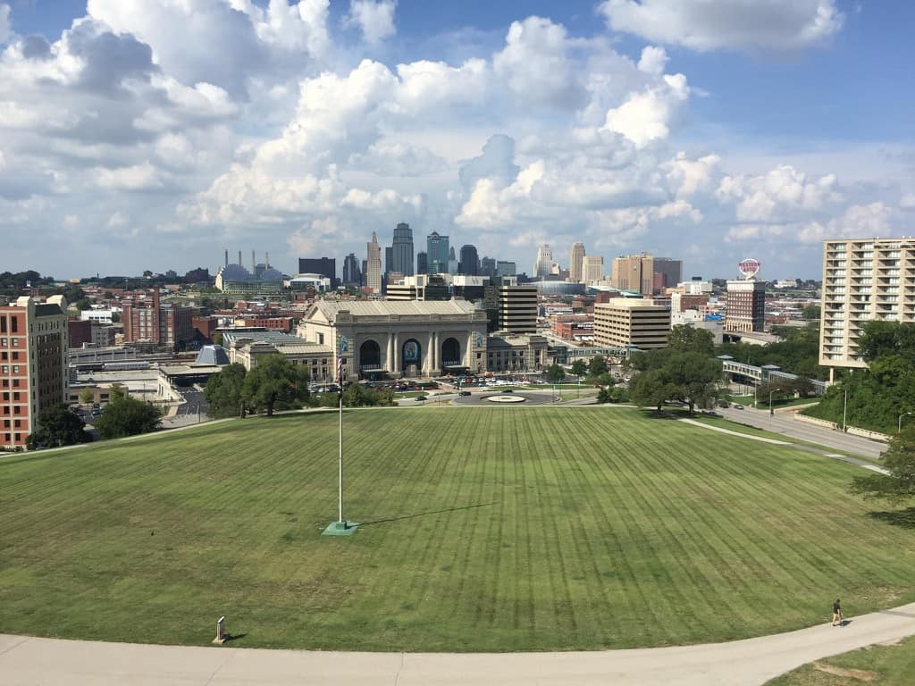 A view of the downtown Kansas City skyline from Liberty Memorial. The bottom half of the picture contains a green lawn. The middle of the picture shows Union Station and numerous city buildings. The top half of the picture contains the sky with many clouds.