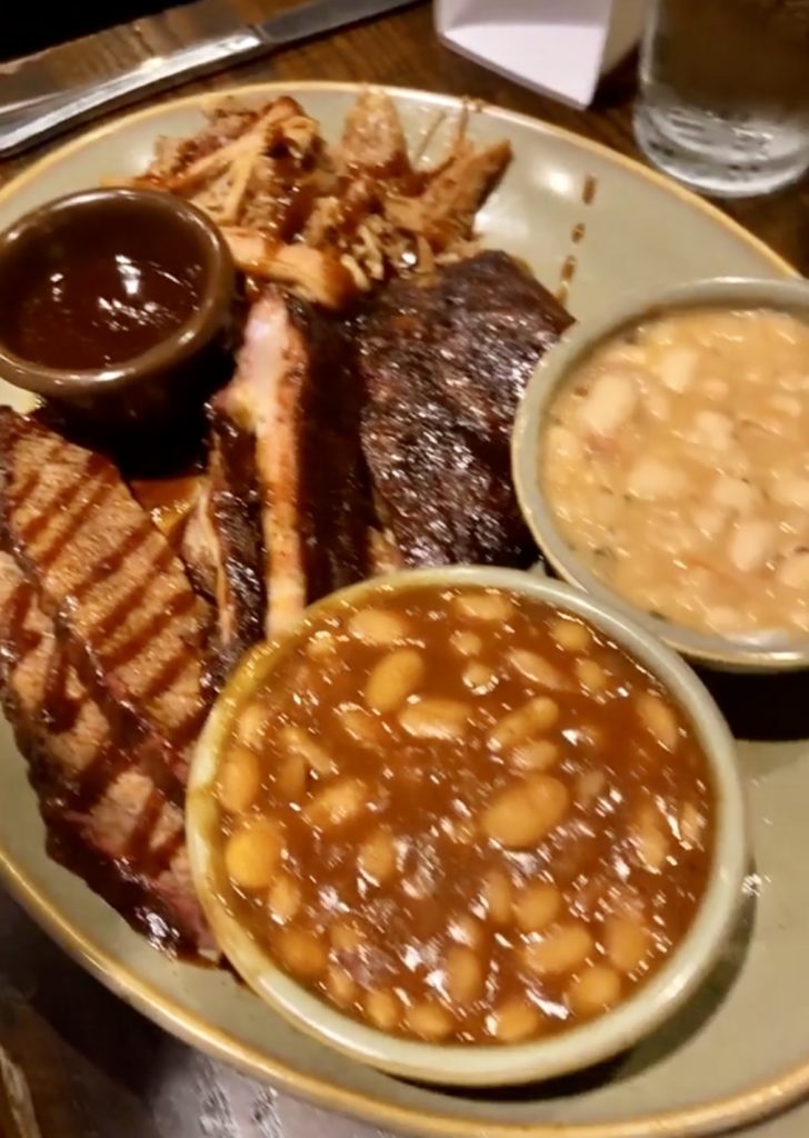 A plate of BBQ meats and various sides from Q39. The meats are ribs, brisket, an chicken, each of which are covered in BBQ sauce. The sides are baked beans and white beans.