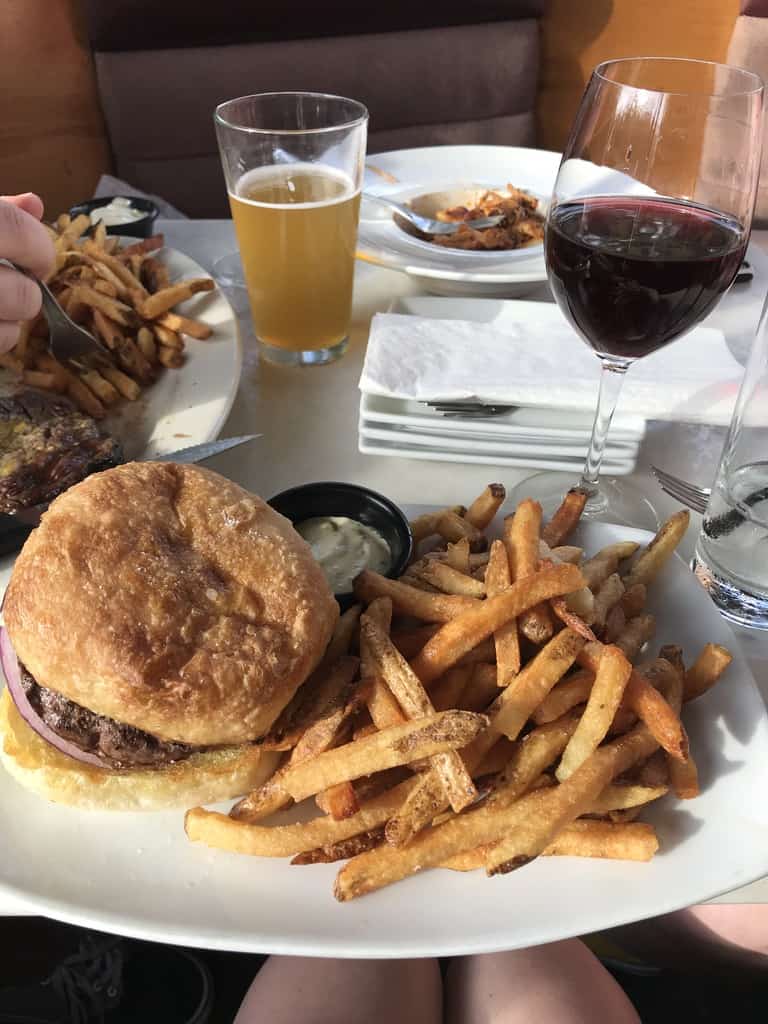 A plate of fries and a burger. A glass of red wine sits behind the plate. A glass of beer and another plate of fries are in the background,.