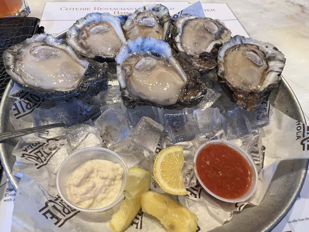 A plate of six raw oysters on ice presented with sides of lemon slices, horseradish, and cocktail sauce.