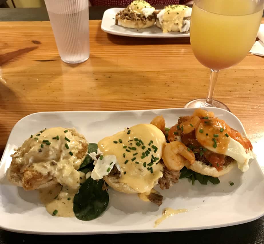 Three egg benedicts, each of a different variety, on a white plate. A mimosa is placed behind the plate.
