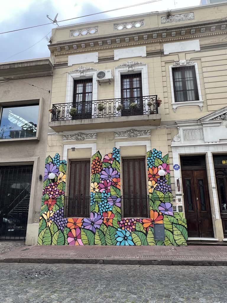 A building with floral street art on the bottom half. The flowers are purple, blue, pink, orange, and yellow and are separated by green leaves.