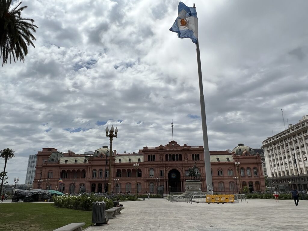 La Casa Rosada (i.e. The Pink House). In front of the pink house is a plaza with a giant Argentina flag and palm trees.