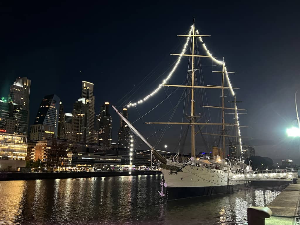 A large white boat with large sails decorated with lights sits on a river. Numerous skyscrapers are shown in the background.