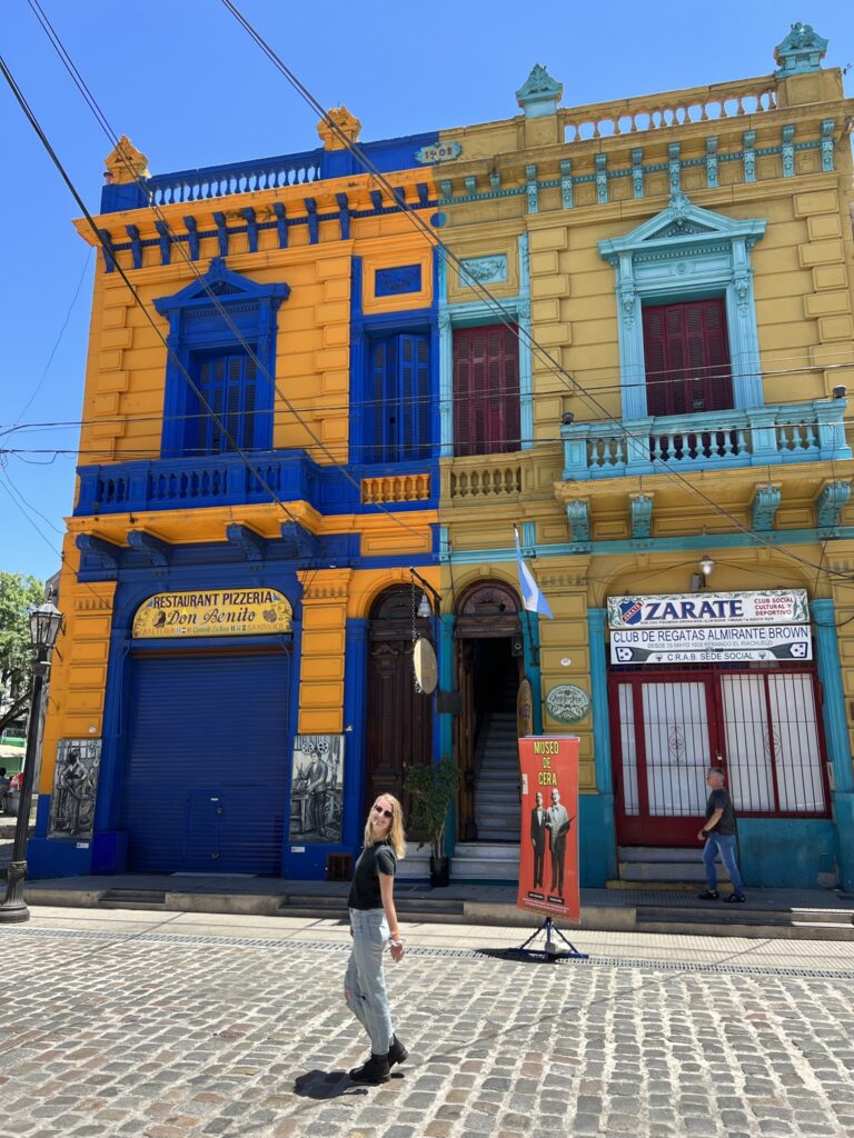 Stephanie (the author of this post) standing in the street in front of two brightly colored buildings. One building is bright orange and blue, and the other building is bright teal and yellow.