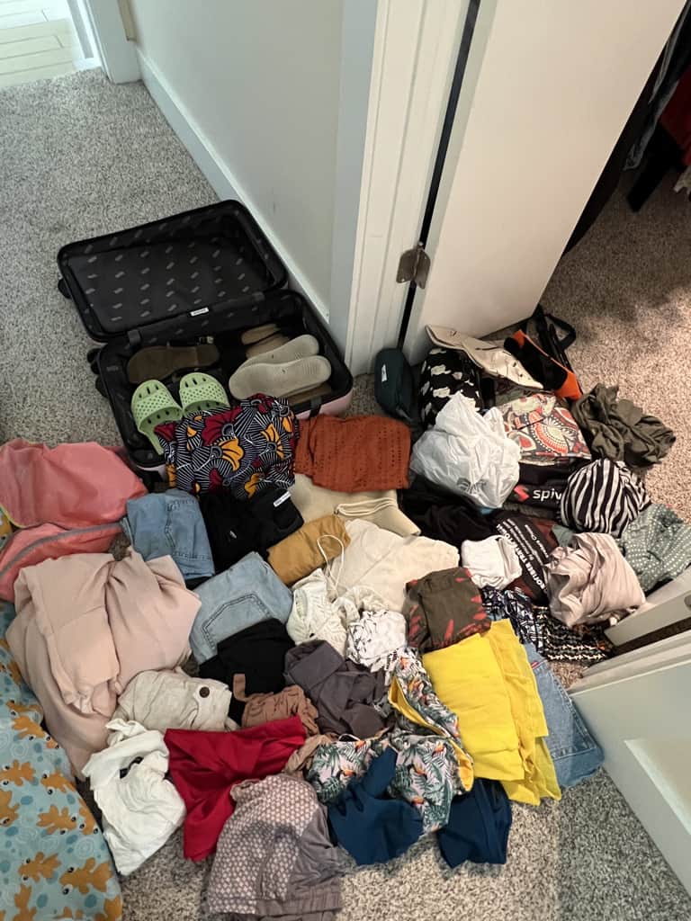 An open suitcase containing four pairs of shoes is sitting beside a large pile of clothes that were formerly packed in the suitcase. 