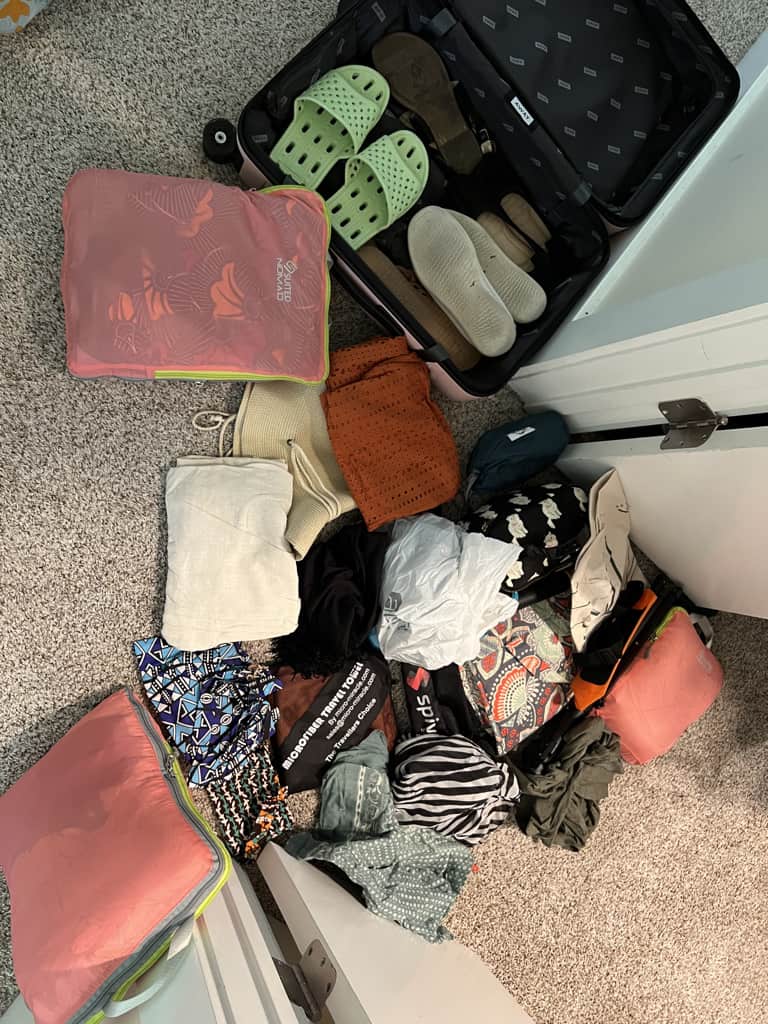 An open suitcase containing four pairs of shoes is sitting beside a pile of clothes that were formerly packed in the suitcase and a few compression packing cubes