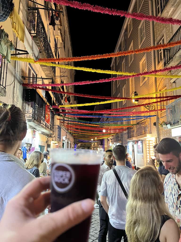 A street in Lisbon at night decorated with streamers and filled with people. Stephanie (the author) is holding up a sangria for a photo.
