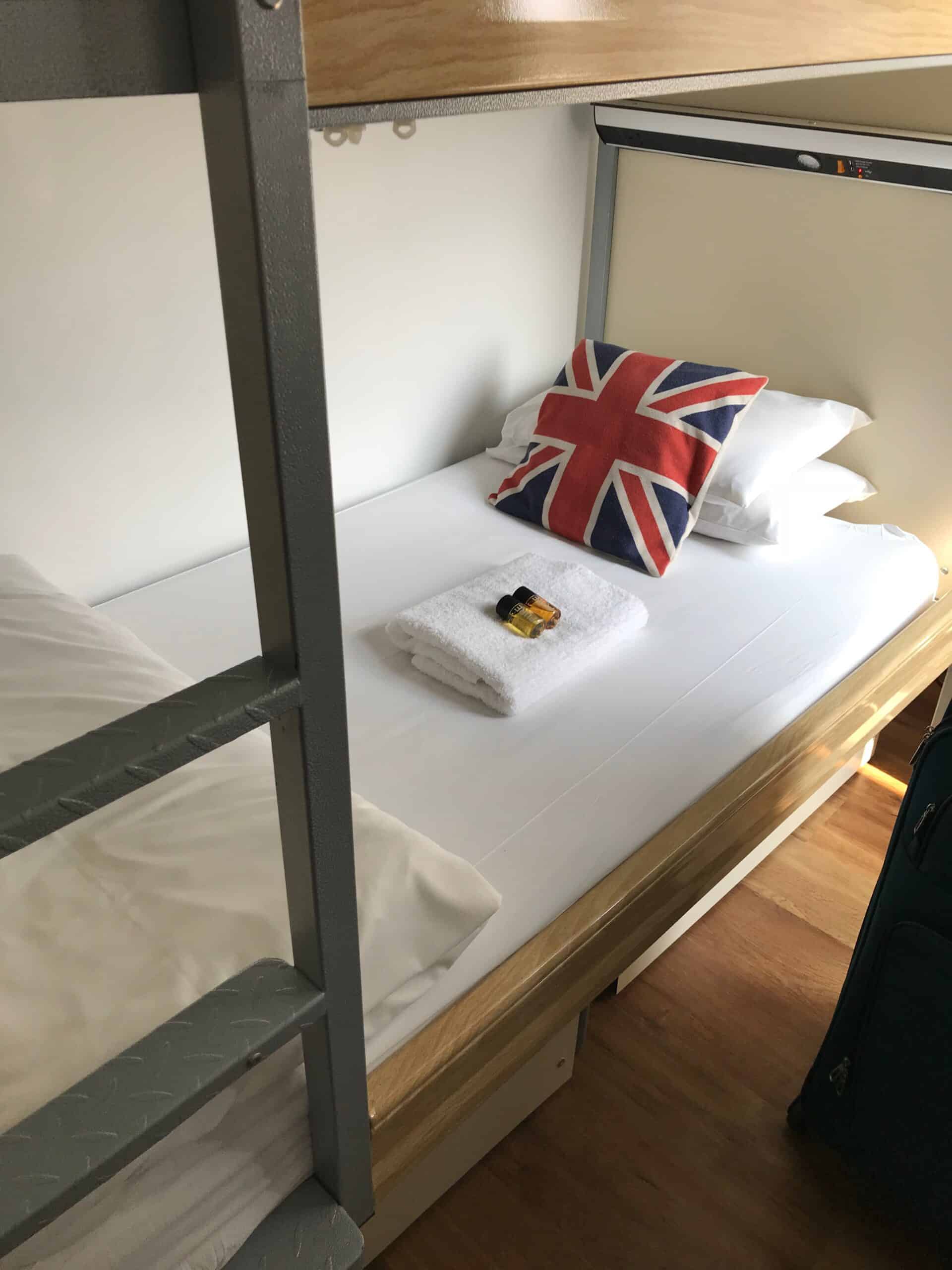The bottom bunk of a bunk bed in a hostel in London. The bed is topped with a blanket, a towel with toiletries, and three pillows. One of the pillows is decorated with the United Kingdom flag.